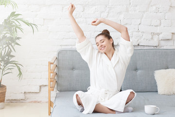Young woman in white bathrobe stretching on couch after wake up in the white loft apartment