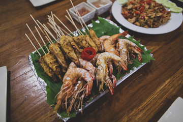 Asian Thai snacks are located on a plate standing on a wooden table. There are shashliks on skewers, fried shrimps and vegetables. The plate is lined with green leaves. Close up.