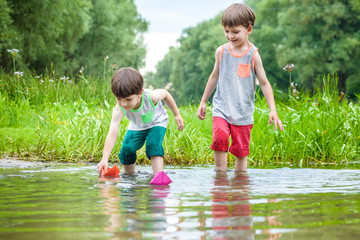 Two little brothers playing with paper boats by a river on warm and sunny summer day.