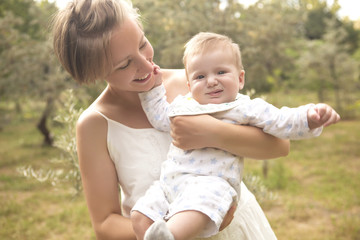 Cute happy caucasian woman with short haircut in white dress in park relaxing with her baby boy son feeling good, smiling. Summer sunset, outdoors. Copy Space