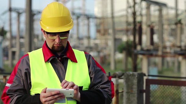 Worker uses cell phone at heat electric power station near outdoor switchgear. Electrician at power plant uses smartphone