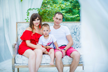 Happy young family is resting on couch in an arbor on background of summer park