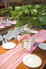 Decoration for a wedding ceremony on a back yard with tables, plates, and vases full of anthurium flowers and monstera leafs. Pink and green colors. Space for text