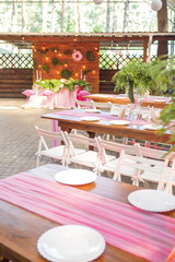 Decoration for a wedding ceremony on a back yard with tables, plates, and vases full of anthurium flowers and monstera leafs. Pink and green colors. Space for text