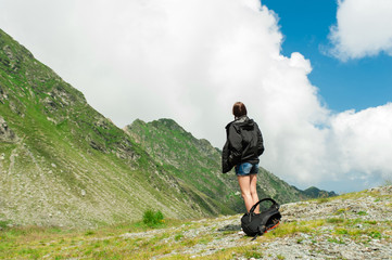 Young lady hiker with backpack sitting on mountain top admiring mountain landscape around and blue...