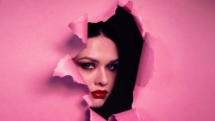 Face beautiful girl with bright make-up, with pouting lips, with red lipstick, appears in torn opening in pink paper background. Creative advertising women's cosmetics. Girl peeks into hole.
