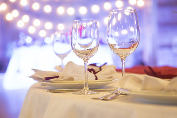 Set of glassess and plates on a wedding table surrounded by light garland and wine color flowers and tablecloth. Interior. Copy space