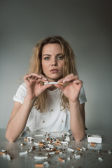 Portrait of serene female holding cigarette and looking at camera, smashed cigars are lying on table. Isolated on background