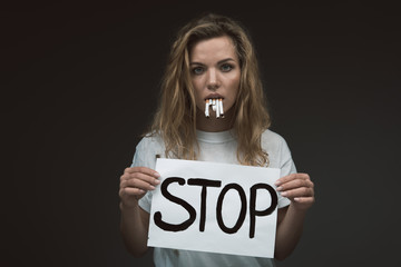 Waist up portrait of severe female person holding cigarettes in mouth and paper with stop word in...