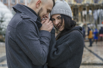Gorgeous beautiful caucasian woman with a man on a walk in european city at winter. Couple wear casual grey outfit and hugging each other. Cold weather, christmas lights on a background