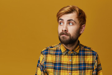 Young hipster curving his lip wears plaid yellow shirt looking aside. Serious expression. Isolated over yellow background with copyspace