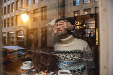 Playful loving couple meeting in cozy cafe. Woman is kissing man with fondness while covering his eyes. View from glass window