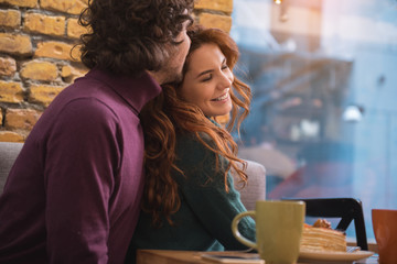 Happy young woman enjoying hug of her husband. They are sitting in cafeteria and smiling. Romantic entertainment concept