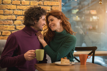Happy young man and woman are flirting and smiling. They are sitting at table in cozy cafeteria and drinking coffee