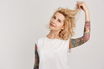 Portrait of young attractive girl wears casual white t-shirt touching her hair. Has tattoos over her arms. isolated over white studio wall