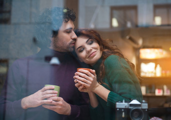 Portrait of joyful woman enjoying hot of her boyfriend. Man is kissing her forehead with gentleness while holding mug of coffee
