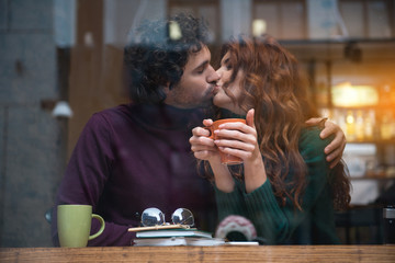 Cheerful young man and woman having romantic kiss in lips. They are sitting at table in cozy cafe