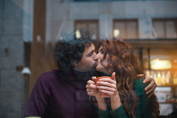Fototapeta premium Happy loving couple kissing with gentleness. They are embracing while girl is holding mug of hot coffee. View from the glass window