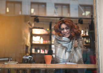 Portrait of cute girl enjoying cozy atmosphere in cafeteria. She is wrapped by warm blanket and smiling. Copy space