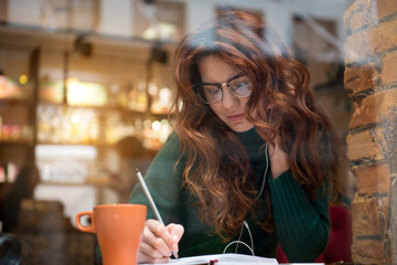 Relaxed young woman is writing into notebook with inspiration. She is listening to music from...