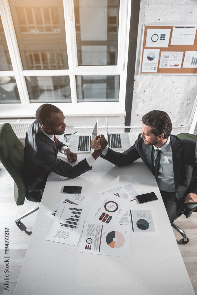 Poster battle of strength. top view of two strong businessmen are sitting at table and competing on arms. t