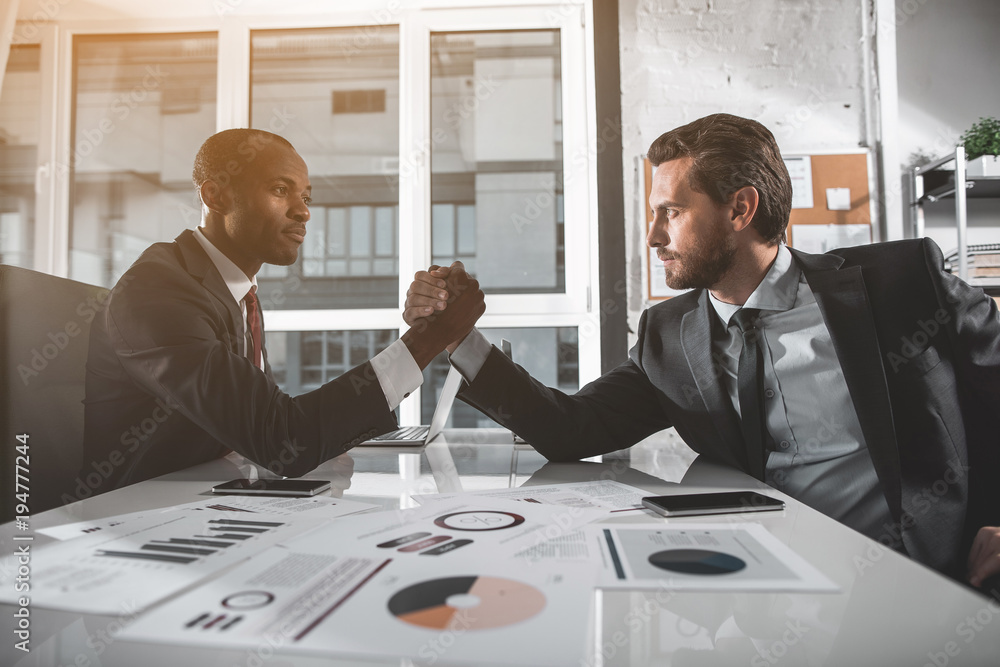 Poster side view profile of two men are sitting opposite with one elbow resting on table and arm wrestling.