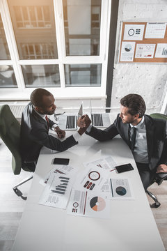 Battle Of Strength. Top View Of Two Strong Businessmen Are Sitting At Table And Competing On Arms. They Are Looking At Each Other Closely While Trying To Force Each Other's Arm Down Onto The Desk