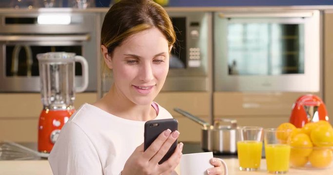 A woman in the kitchen while having breakfast and sipping tea or milk in the cup sends a message or calls with the phone and smiles. Concept of: social network, message, technology.