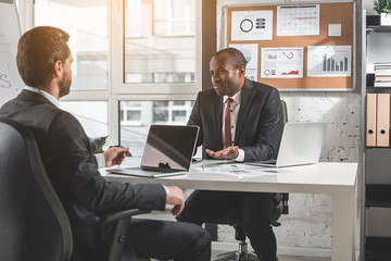 Listen attentively. Concentrated young businessmen in suit are sitting at table with laptops while...