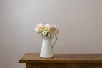 White dahlias in jug on wooden table against neutral wall background