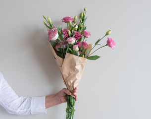 Woman's hand holding bouquet of pink lisianthus flowers wrapped in in brown paper against neutral...
