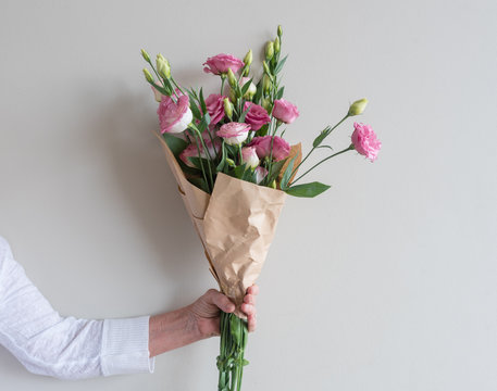 Woman's Hand Holding Bouquet Of Pink Lisianthus Flowers Wrapped In In Brown Paper Against Neutral Wall Background