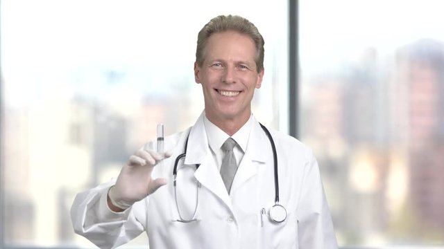 Smiling doctor with syringe, blurred background. Mature professional doctor holding syringe on blurred background.