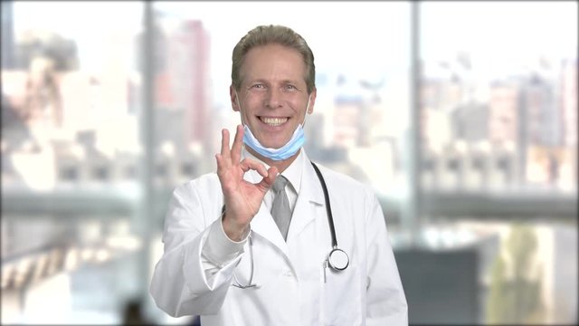 Pleased doctor with ok gesture. Handsome mature doctor showing ok sign and looking at camera, window city background.