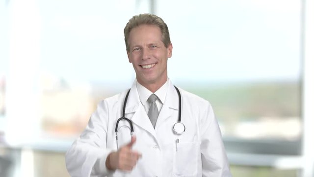 Happy male cardiologist on blurred background. Cheerful male doctor with stethoscope gesturing thumb up on blurred background.