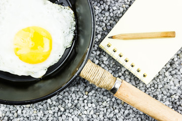 Egg in a frying pan and a notebook on gray gravel. Cooking recipe, homemade lunch.