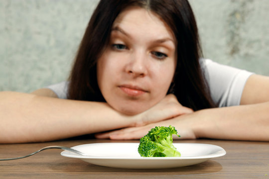 sad young brunette woman dealing with anorexia nervosa or bulimia having small green vegetable on plate. Dieting problems, eating disorder.