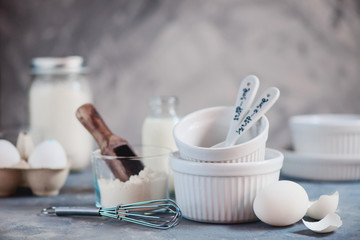 Baking cocnept with a stack of porcelain baking forms, , measuring spoons, eggs, flour, and milk on a concrete background. High key food photography with copy space.