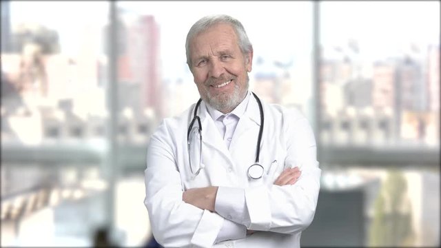 Cheerful caucasian doctor crossed arms. Elderly male doctor smiling and looking at camera. Happy medical doctor folded arms on blurred background.