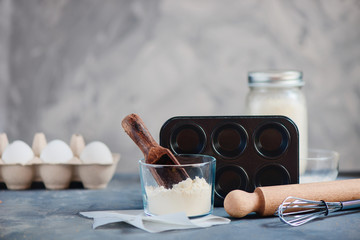 Cooking concept with baking tools and ingredients. Flour scoop, muffin tin, a whisk, a rolling pin, milk and eggs on a concrete background. High key food photography with copy space.
