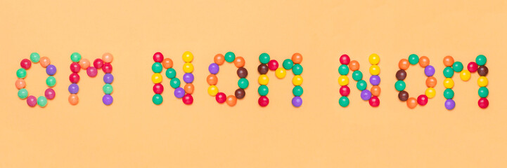 The word OM NOM NOM is written by sweet colorful chocolate candy on pink background