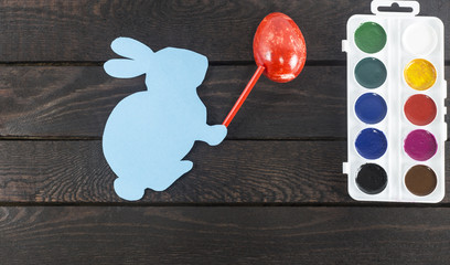 card bunny with marker on it's hand, painting egg into red on dark brown wooden background.