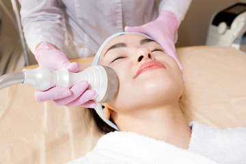 Obraz na płótnie Canvas Headshot of pretty young woman wearing bathrobe and headband lying on treatment table of beauty salon while getting ultrasound face cleaning