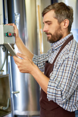 Profile view of bearded middle-aged brewer wearing apron using digital tablet while trying to repair equipment at modern beer factory