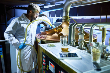 Confident middle-aged worker of modern brewing plant wearing white coat and protective goggles...