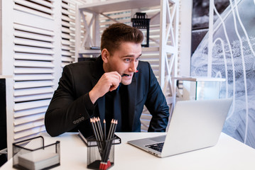 Businessman working behind a laptop, surprised and holding a pencil in his mouth..