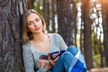 Beautiful blond girl resting in spring or autumn forest read book and sitting with cup of tea and book. Confident caucasian young woman in casual clothes relaxing near big tree in park and holding