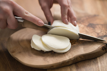 female teen hand slicing mozzarella cheese with knife on wooden board