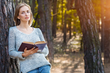 Obraz premium Beautiful blond girl resting in spring or autumn forest read book and standing. Confident caucasian young woman in casual clothes relaxing near big tree in park and holding book. Attractive female