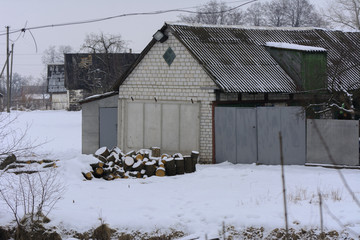 A village dwelling house with outbuildings fenced with a stone fence against a background of white snow in the winter.